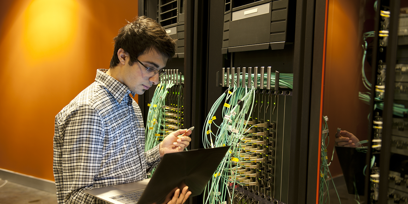 A man with a laptop in front of a network server rack