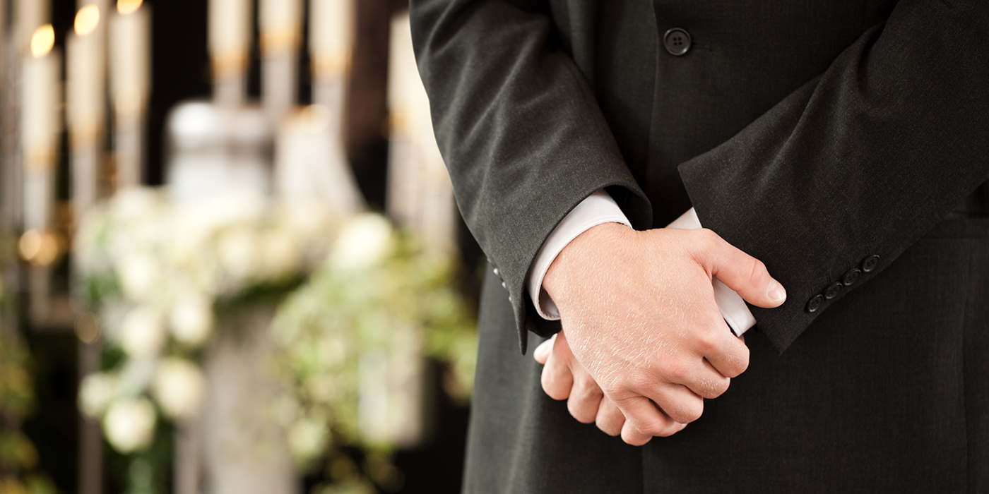 Close up of a person in a suit with crossed hands in front of a flower arrangement