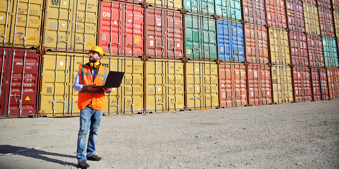 A man in a safety vest and hard hat standing in front of stacks of shipping containers
